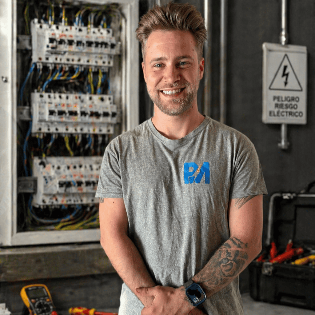 Un técnico sonriente y tatuado posa frente a un cuadro eléctrico abierto. Al fondo se observa una señal de peligro eléctrico junto a equipos de mantenimiento preventivo, mientras que en su camiseta destaca el logotipo de Instalaciones Industriales PM.