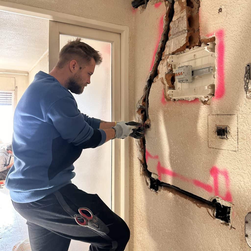 Una persona que lleva guantes está trabajando en el cableado eléctrico a la vista dentro de una pared, en la que se ven una caja de fusibles y marcas pintadas con spray.